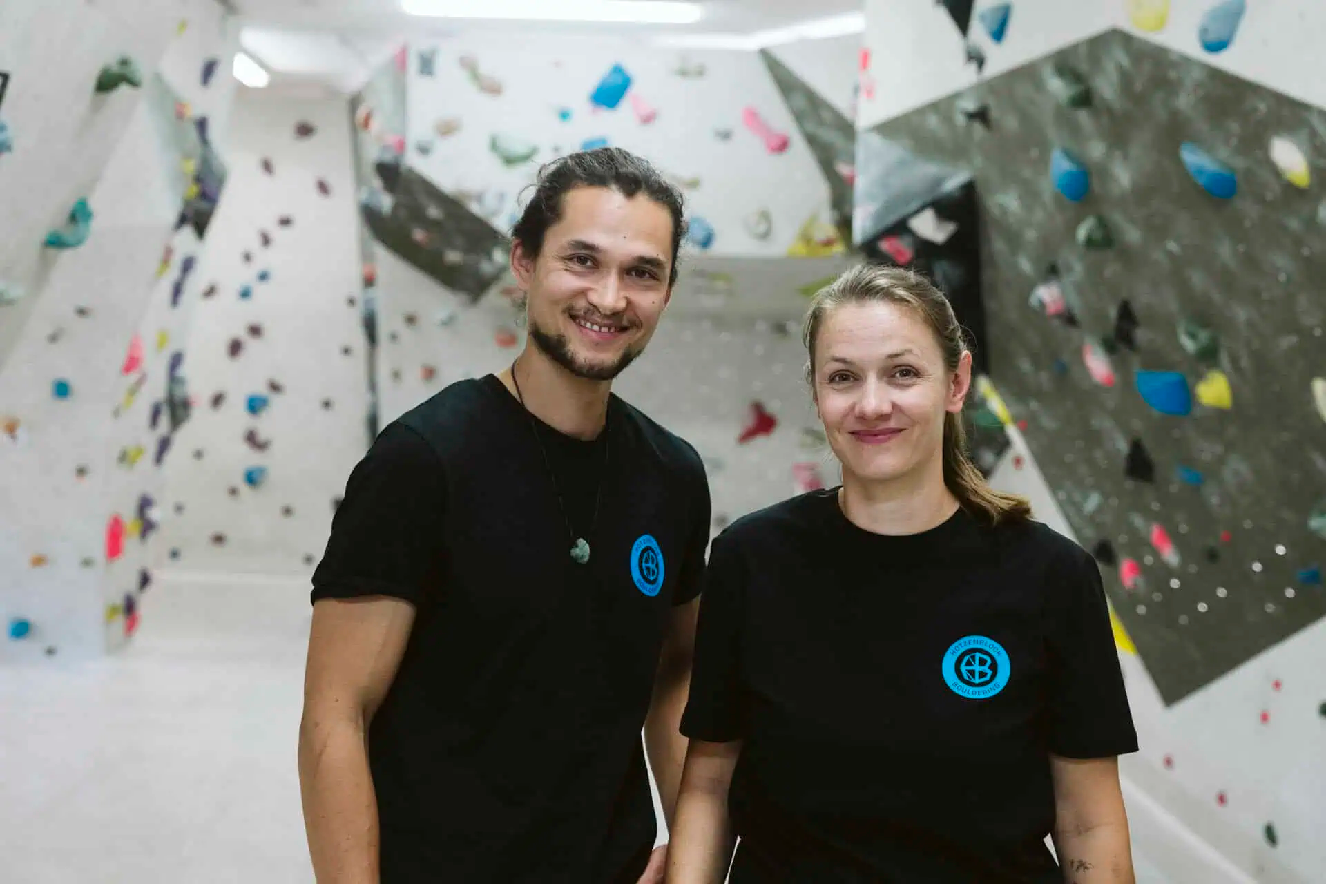 Zwei Personen in sportlicher Kleidung stehen in einer modernen Kletterhalle mit bunten Klettermaterialien im Hintergrund.